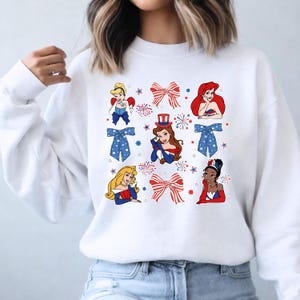 May include: White sweatshirt with cartoon images of princesses, red and blue bows, and fireworks. The design uses patriotic colours and imagery, ideal for celebrating a holiday.