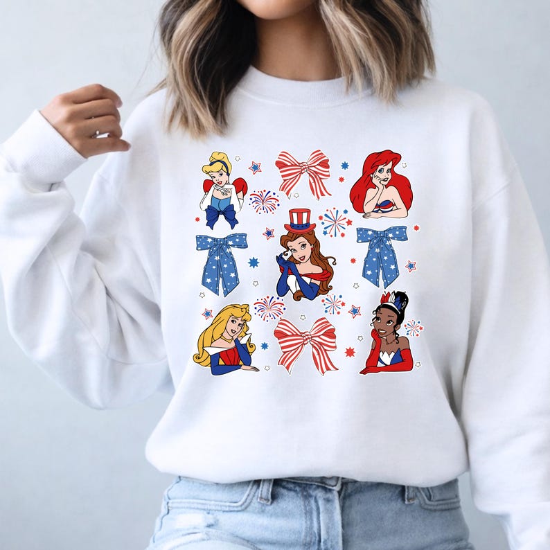 May include: White sweatshirt with cartoon images of princesses, red and blue bows, and fireworks. The design uses patriotic colours and imagery, ideal for celebrating a holiday.