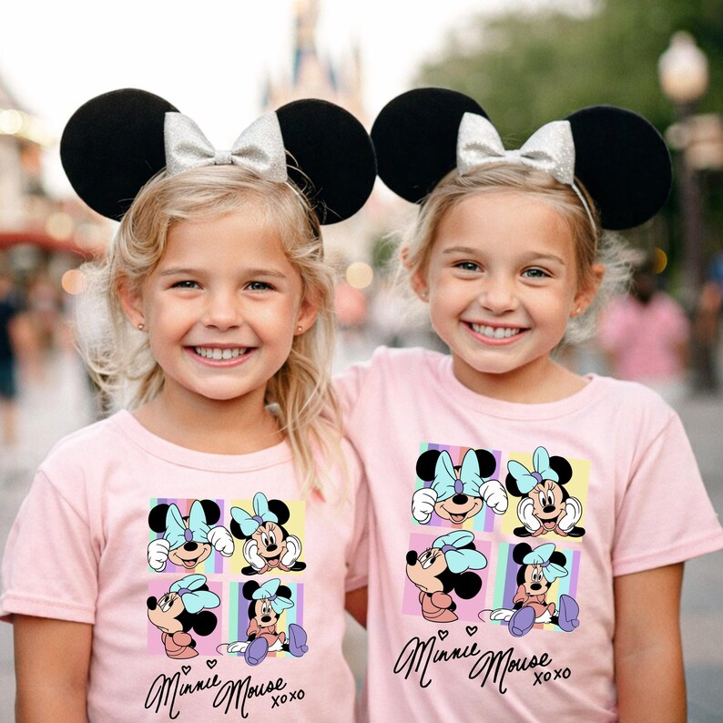 Pu&ograve; includere: Due bambine con orecchie da topo nere con fiocchi argentati e magliette rosa. Le magliette presentano una grafica di Minnie Mouse con la scritta "Minnie Mouse xoxo". Lo sfondo &egrave; sfocato.