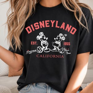 Puede incluir: Camiseta negra con un gr&aacute;fico rojo desgastado de "Disneyland" sobre ilustraciones blancas de Mickey y Minnie Mouse. La camiseta incluye el texto "Est. 1955" y "California", junto con las firmas "Mickey Mouse" y "Minnie Mouse".