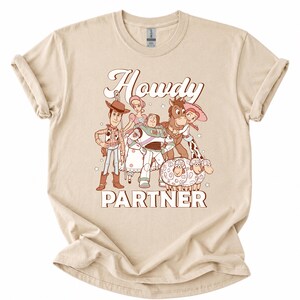 May include: Beige t-shirt featuring a cartoon graphic of characters from a popular animated movie. The graphic includes the text "Howdy Partner" in a retro font. The shirt is made of a soft material and has rolled-up sleeves.
