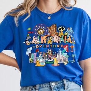 May include: A royal blue t-shirt with a vibrant graphic that reads "CALIFORNIA ADVENTURE". The design incorporates cartoon characters and theme park landmarks, including a Ferris wheel and a castle.