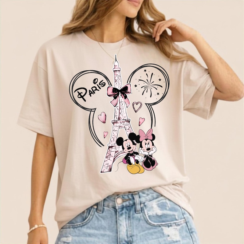 K&ouml;nnte beinhalten: Beiges T-Shirt mit einem Aufdruck des Eiffelturms, Mickey und Minnie Mouse und dem Wort "Paris". Das Design umfasst Herzen, Feuerwerk und eine Schleife. Das Shirt hat eine helle Farbe.