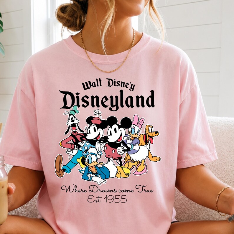 Pu&ograve; includere: T-shirt rosa chiaro con una grafica dei personaggi Disney, tra cui Topolino, Minnie, Paperino, Pippo, Paperina e Pluto. La maglietta presenta la scritta "Walt Disney's Disneyland" e "Where Dreams come True Est. 1955."