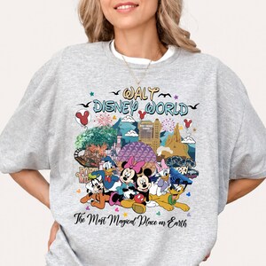 K&ouml;nnte beinhalten: Ein hellgraues Sweatshirt mit einer farbenfrohen Grafik von Disney-Figuren und Wahrzeichen. Das Design zeigt Micky und Minnie Maus, Donald Duck und Goofy, mit dem Text "Walt Disney World" und "The Most Magical Place on Earth."