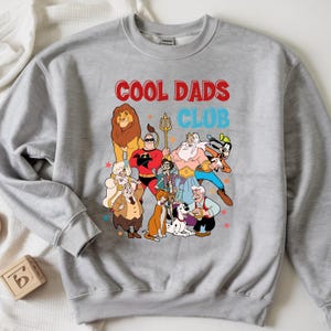 Pode incluir: Um moletom cinza com o texto "COOL DADS CLUB" em vermelho e azul. O gr&aacute;fico apresenta personagens de desenhos animados, incluindo um le&atilde;o e um super-her&oacute;i. O design &eacute; impresso na frente do moletom.