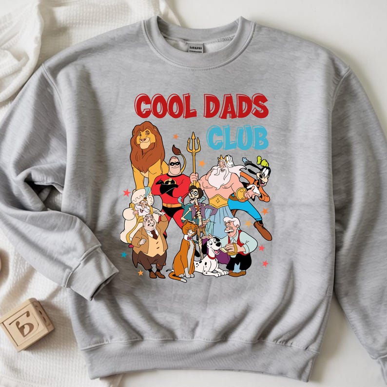 Pode incluir: Um moletom cinza com o texto "COOL DADS CLUB" em vermelho e azul. O gr&aacute;fico apresenta personagens de desenhos animados, incluindo um le&atilde;o e um super-her&oacute;i. O design &eacute; impresso na frente do moletom.