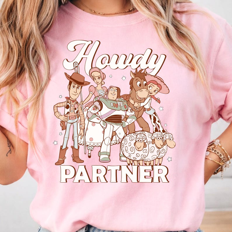 May include: Pink t-shirt featuring a cartoon graphic of characters from a popular animated film. The design includes the word "Howdy" above the characters and the word "Partner" below. The graphic is in shades of brown, white, and blue.