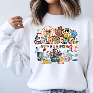 May include: White sweatshirt with a vibrant graphic of Disneyland characters and attractions. The design includes the words "CALIFORNIA ADVENTURE" in a colourful font, surrounded by iconic imagery like Mickey Mouse and a Ferris wheel.