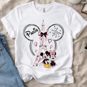 K&ouml;nnte beinhalten: Wei&szlig;es T-Shirt mit einem Aufdruck des Eiffelturms, einer rosa Schleife, Herzen und dem Wort "Paris". Micky und Minnie Maus sind abgebildet. Das Design wird von Micky-Maus-Ohren und Feuerwerk umrahmt.