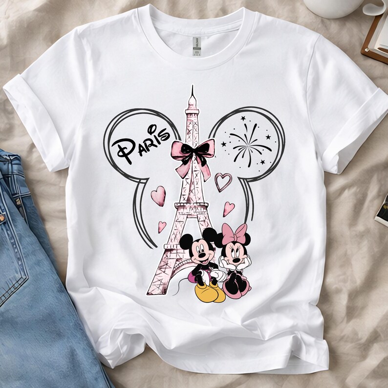 K&ouml;nnte beinhalten: Wei&szlig;es T-Shirt mit einem Aufdruck des Eiffelturms, einer rosa Schleife, Herzen und dem Wort "Paris". Micky und Minnie Maus sind abgebildet. Das Design wird von Micky-Maus-Ohren und Feuerwerk umrahmt.