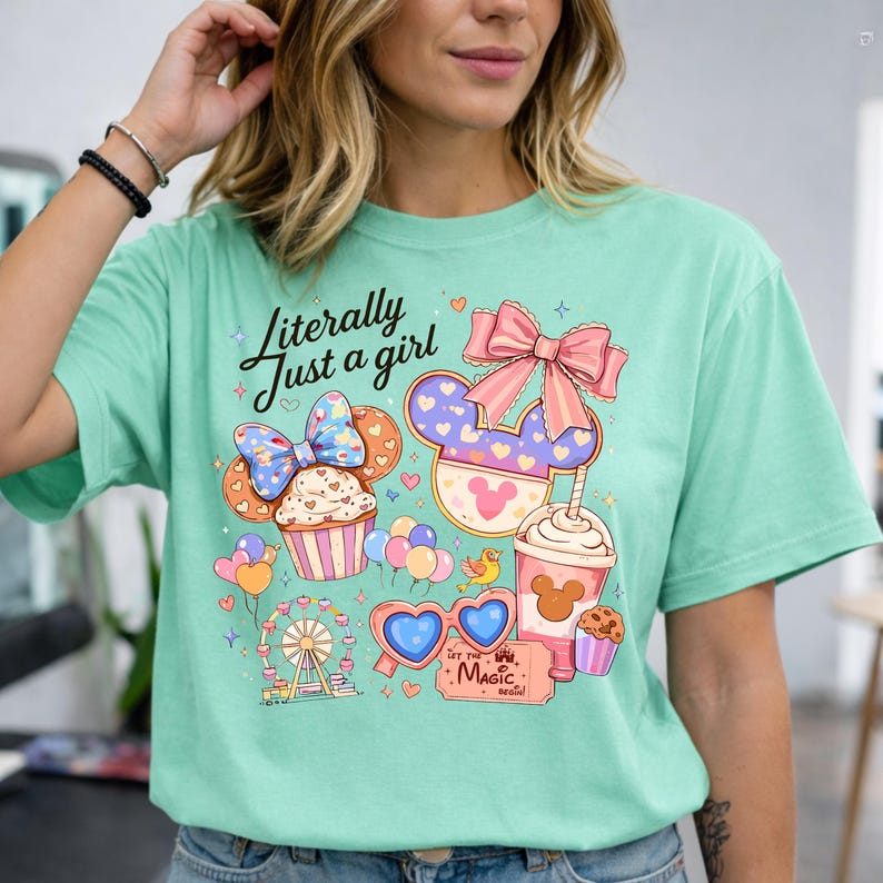 Pode incluir: Camiseta verde menta com um gr&aacute;fico colorido com um cupcake, orelhas do Mickey Mouse, uma bebida e o texto "Literally Just a girl". O design inclui uma roda gigante, &oacute;culos de sol em forma de cora&ccedil;&atilde;o e as palavras "Let the Magic Begin".