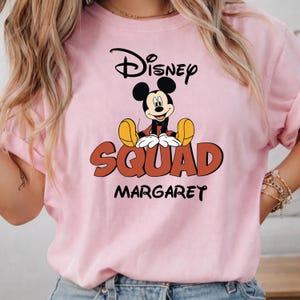 Op de afbeelding: Lichtroze T-shirt met een Mickey Mouse-ontwerp. Het Disney-logo staat bovenaan, met "SQUAD" in grote oranje letters onder Mickey. De naam "MARGARET" staat onder het woord "SQUAD" afgedrukt.
