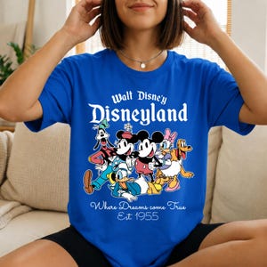 Pu&ograve; includere: T-shirt blu reale con una grafica dei personaggi Disney, tra cui Topolino e Minnie, Paperino, Pippo e Paperina. Il testo "Walt Disney's Disneyland" e "Where Dreams come True Est. 1955" sono stampati sulla maglietta.