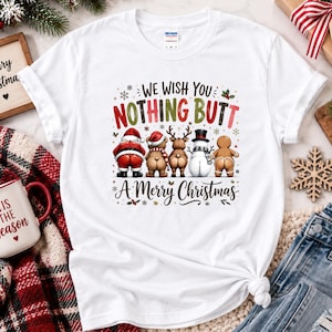 May include: A white short-sleeve t-shirt featuring a graphic of Santa Claus, a bear, a reindeer, a snowman, and a gingerbread person facing away. The text reads: WE WISH YOU NOTHING BUTT A MERRY CHRISTMAS, decorated with small snowflakes and holly.