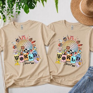 May include: Two beige t-shirts with a colourful graphic featuring Mickey Mouse, Donald Duck, and other characters. The graphic includes the text "WORLD Tour 2026" and various landmarks.