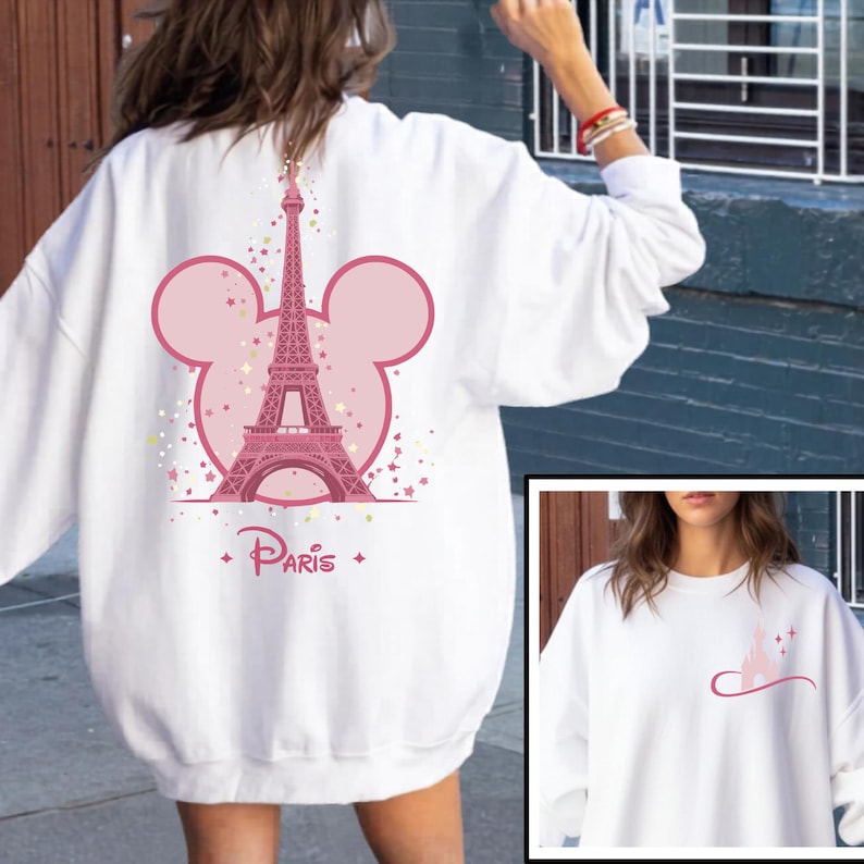 May include: White sweatshirt featuring a pink Eiffel Tower design within a Mickey Mouse ear outline. The word "Paris" is written in a stylized font below the tower. The design includes stars and confetti.