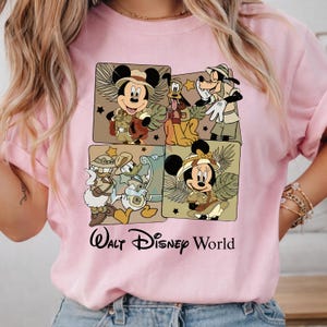Puede incluir: Sudadera rosa claro con un gr&aacute;fico de Mickey Mouse, Pluto, Goofy, Donald y Minnie Mouse con trajes de safari. Las palabras "Walt Disney World" est&aacute;n impresas en negro debajo. La sudadera tiene mangas largas.