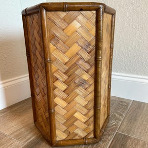 Peut inclure: Une corbeille à papier hexagonale construite avec un cadre en bambou brun foncé et des panneaux en rotin tressé beige clair. La structure présente des tiges de bambou verticales à chaque coin et une bordure en bambou horizontal.
