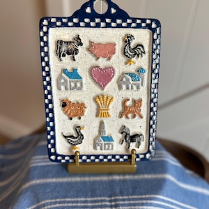 Könnte beinhalten: Ein rechteckiges Wandbild aus Keramik mit einem dunkelblau-weißen Karorand. Die cremefarbene, gesprenkelte Oberfläche zeigt zwölf erhabene, bunte Symbole, darunter eine Kuh, ein Schwein, ein Hahn, ein Herz, ein Schaf, Weizen, eine Katze, eine Gans und Gebäude.