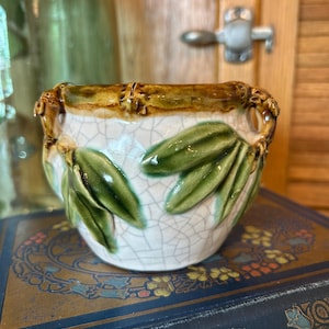 May include: A small ceramic planter with a white crackle glaze finish, featuring raised green leaf motifs and a brown bamboo-textured rim. The vessel has two small handles on the sides and a rounded body shape with a glossy, textured surface.