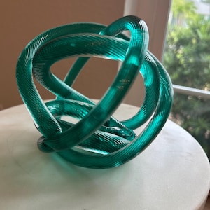 May include: A translucent teal glass sculpture shaped into a continuous, knotted loop. The object features a ribbed, linear texture running along the length of the glass tubing, creating a complex, intertwined form that catches the light.