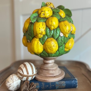 May include: A decorative topiary sculpture shaped like a lemon tree with bright yellow lemons and green leaves, mounted on a pedestal base. Beside it are two spherical objects with white and brown stripes and a small, dried brown seed pod.