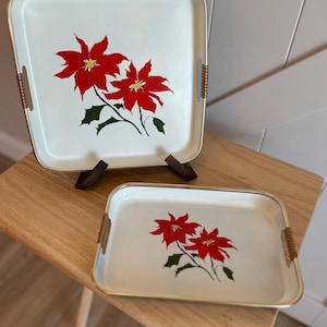 May include: Two off-white metal serving trays with rounded corners and gold-tone rims. Each tray features a central graphic of two red poinsettia flowers with green leaves. The side handles are wrapped in a textured, light brown material.