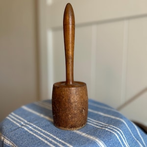 Könnte beinhalten: Ein hölzerner Vintage-Schlägel mit einem zylindrischen Kopf, der eine verwitterte, strukturierte Oberfläche aufweist, und einem langen, glatten, konischen Griff. Das Werkzeug ist aus braunem Holz gefertigt und zeigt natürliche Maserungen.
