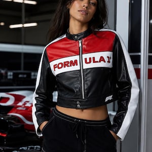 Könnte beinhalten: Eine bauchfreie Lederjacke in Schwarz, Rot und Weiß mit Stehkragen, durchgehendem silbernem Reißverschluss und dem in Rot auf die Brust gedruckten Schriftzug "FORMULA 1". Die Ärmel haben weiße Einsätze und die Jacke zeigt ein kleines F1-Logo.