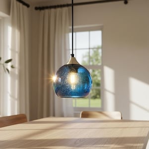 May include: A blue glass pendant light with a speckled pattern hangs above a wooden table. The light fixture has a gold-colored top and a visible lightbulb. The room has large windows and wooden chairs.