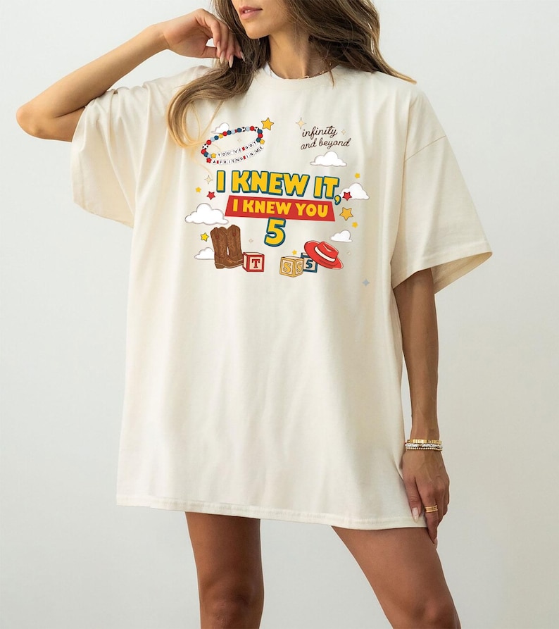 May include: An off-white short-sleeve t-shirt featuring a graphic print with the text "I KNEW IT, I KNEW YOU 5" and "infinity and beyond". The design includes illustrations of brown cowboy boots, a red hat, alphabet blocks, and colorful stars.