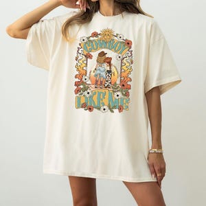 Puede incluir: Una camiseta de manga corta color hueso con un estampado gr&aacute;fico de dos figuras con sombreros de vaquero y botas. El dise&ntilde;o incluye un sol, flores y llamas, con el texto COWBOY LIKE ME impreso en letras grandes de color azul.