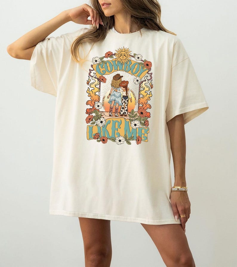 Puede incluir: Una camiseta de manga corta color hueso con un estampado gr&aacute;fico de dos figuras con sombreros de vaquero y botas. El dise&ntilde;o incluye un sol, flores y llamas, con el texto COWBOY LIKE ME impreso en letras grandes de color azul.
