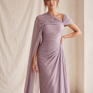 May include: A lavender-colored, one-shoulder formal gown with a draped cape detail. The dress features a fitted bodice with ruching and a flowing skirt. A gold necklace is visible. The fabric appears to be a soft, lightweight material, suitable for special occasions.