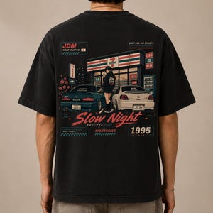 May include: A black short-sleeve t-shirt featuring a graphic print on the back. The design depicts a convenience store at night with a teal car, a white car, and a person standing between them. Text includes JDM, Slow Night, and 1995.