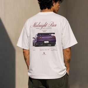 Peut inclure: Un t-shirt blanc à manches courtes avec un imprimé au dos représentant une voiture de sport violette. Le texte indique Midnight Run, Where Assets Appreciate, Night Run Co. Owners Club, accompagné de caractères japonais et d'un logo rouge.