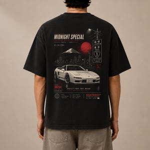 Peut inclure: Un t-shirt noir à manches courtes avec une impression graphique au dos. Le motif représente une voiture de sport blanche, un soleil rouge, une silhouette de montagne et du texte japonais. On y lit "MIDNIGHT SPECIAL" et "BUILT FOR THE ROAD."