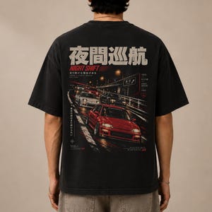 Op de afbeelding: Een zwart t-shirt met korte mouwen en een grote afbeelding op de achterkant. Het ontwerp toont een rode auto die 's nachts op een snelweg rijdt, met Japanse tekst, de woorden NIGHT SHIFT en diverse kleine tekstuele elementen in wit en rood.