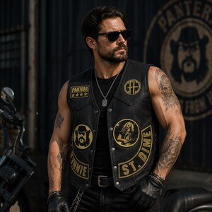 May include: A black leather vest featuring gold embroidered patches, including the text Pantera, Vinnie, and St. Dime, along with circular portraits and a logo. The vest has a button-front closure and is worn over a black shirt with a silver necklace.