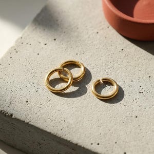 May include: Three gold-colored hoop earrings, two interlocked, and one separate, rest on a gray concrete surface. A small, terracotta-colored bowl is in the background. The earrings are simple and minimalist in design.
