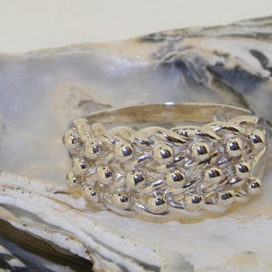 May include: A silver ring with a textured design featuring small, round, raised elements. The ring is set against a neutral background, highlighting its polished surface and intricate details. The ring's design is reminiscent of a woven pattern.
