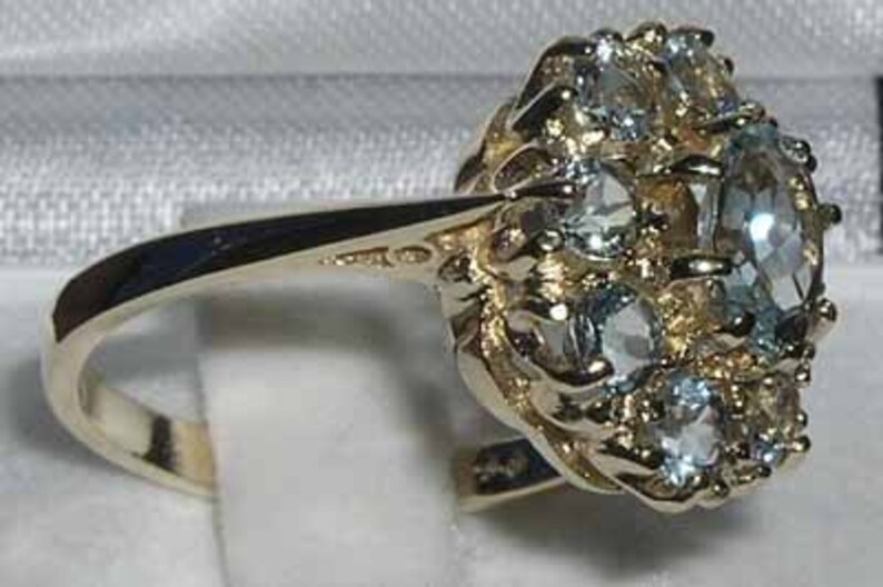 May include: A gold ring featuring a cluster of light blue gemstones. The ring's band is a polished gold, and the gemstones are set in a detailed, textured gold setting. The ring is displayed on a white surface.