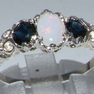 May include: A silver ring with an oval opal centre stone and two dark blue sapphires. The ring has a decorative, textured band with small accent stones. The gemstones are set in a detailed, ornate setting.