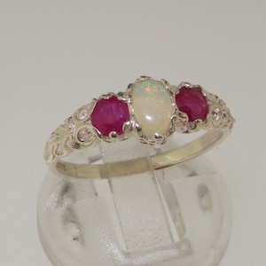 Op de afbeelding: Zilveren ring met een ovale opaal en twee robijnen. De ring heeft een gedetailleerd, bloemachtig ontwerp met kleine diamanten. De edelstenen zijn gezet in een klauwzetting.