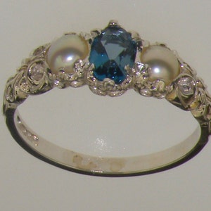 May include: A silver ring with a blue topaz gemstone and two white pearls. The ring has a delicate, ornate design.