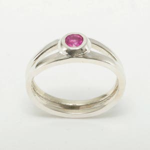 May include: A silver ring with a pink gemstone set in a bezel setting. The ring has a double band design.
