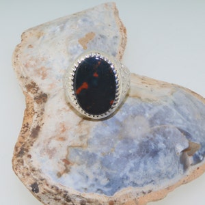 May include: A silver ring with an oval-shaped black and red gemstone set in a silver bezel. The ring is on a white and gray background.