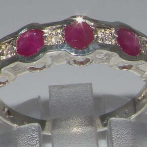 May include: A silver ring with a row of oval-cut, bright red gemstones and smaller clear stones. The ring's band is polished, and the gemstones are set across the top. The ring is a piece of jewellery.