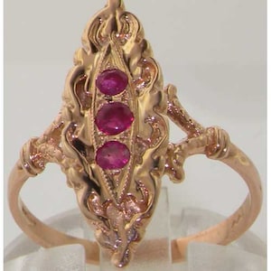 May include: A gold-toned ring featuring three oval-cut, deep red gemstones set vertically in an ornate, filigree design. The ring has a detailed, vintage aesthetic with a delicate band and decorative flourishes.